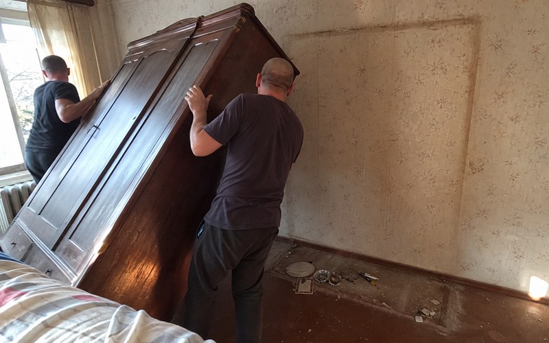 Two workers tilting a tall wooden wardrobe to dismantle it
